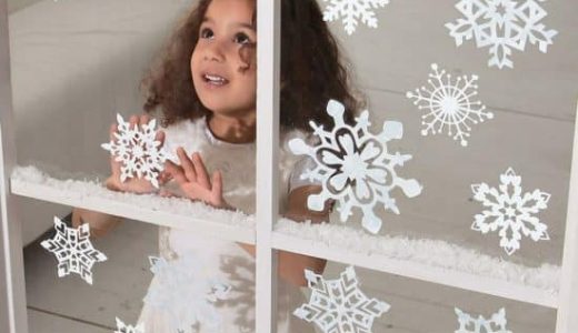 A little girl behind a window pasted with paper Christmas cutouts in the shape of snowflakes